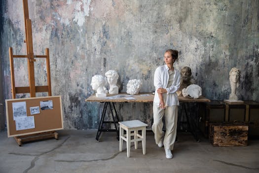 Femme contemplant des sculptures sur une table dans un atelier artistique avec des croquis et un chevalet en arrière-plan.